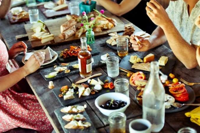 Overhead shot of a wooden table laden with various food items like cheese, bread, and fruit, with several people seated around it.