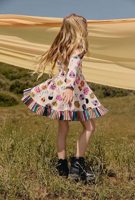 Young girl with blonde hair poses in a dress printed with pumpkins and ghosts, wearing black boots, standing in a grassy field.