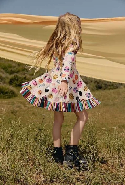 Young girl with blonde hair poses in a dress printed with pumpkins and ghosts, wearing black boots, standing in a grassy field.