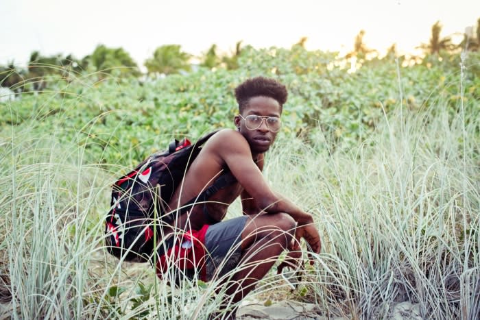 Young man with backpack crouches in beach grass near trees, wearing glasses and shorts in a lifestyle portrait under bright sky.