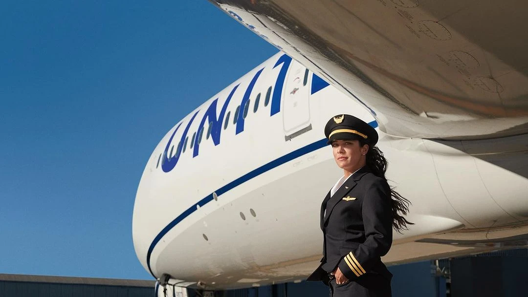 Female pilot in uniform stands confidently in front of a large commercial airplane, under a clear blue sky, in an outdoor setting.
