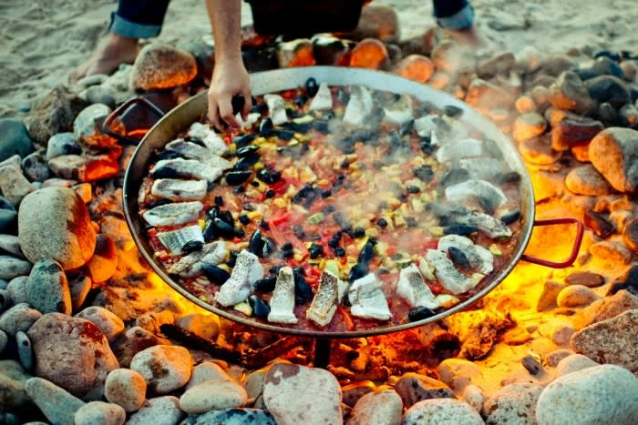 Large paella pan cooking over an open fire on a sandy beach surrounded by smooth stones, with a person's hands and feet visible.