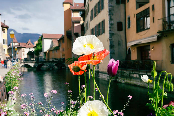 Scenic view of Annecy canal with historic buildings and vibrant flowers in the foreground on a partly cloudy day, creating a serene ambiance.