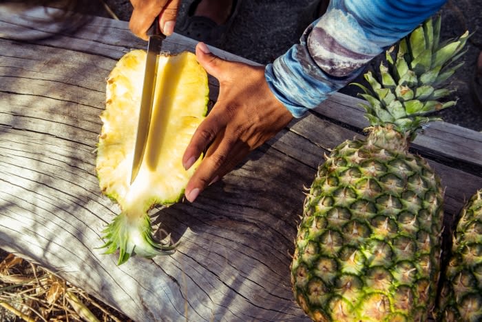 Person cutting a pineapple in half with a knife on a rustic wooden surface, close-up, showcasing the yellow fruit and green leaves.