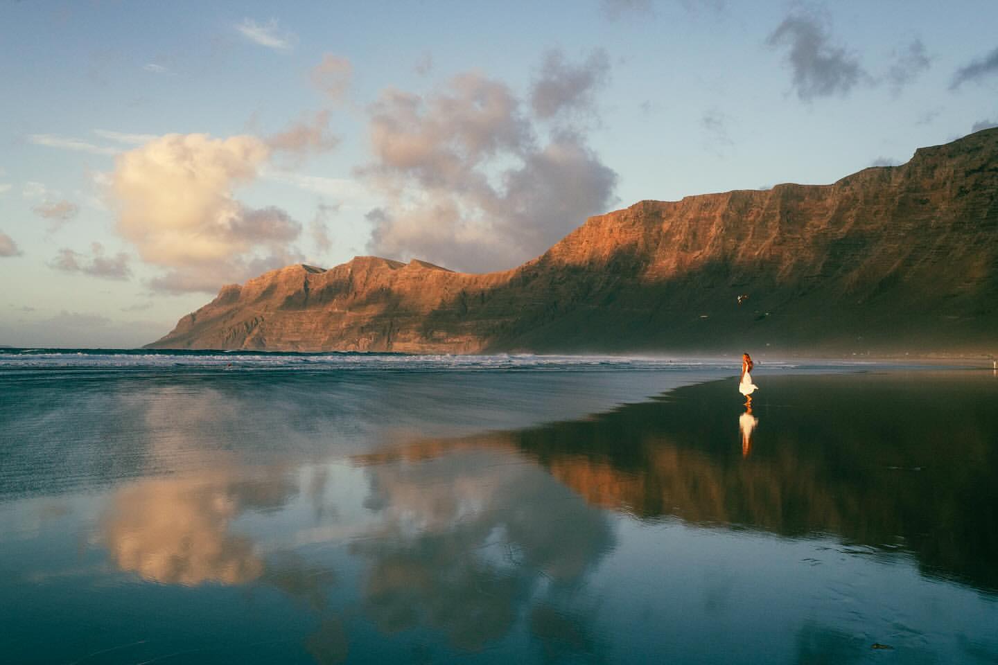 Woman in white dress walking on wet sandy beach near mountains under a blue sky with clouds during daytime.