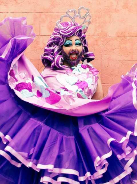 Person in drag with purple wig, beard and dress poses in front of a brick wall. The makeup is dramatic, and expression is excited.