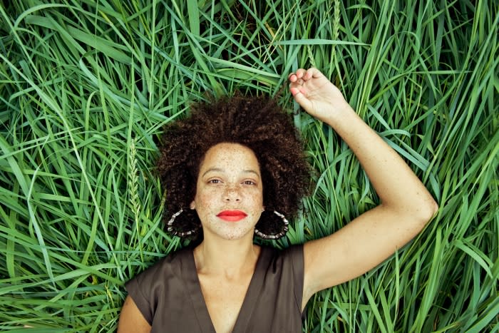 Woman with curly hair and red lipstick lying in green grass outdoors, wearing black top and hoop earrings in natural sunlight.