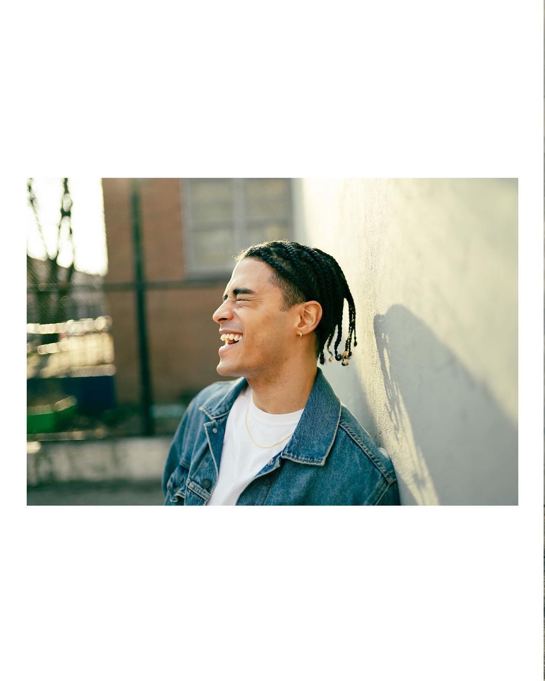 Portrait of a laughing man in a denim jacket leaning against a light wall outdoors, photographed by Gabriela Herman.