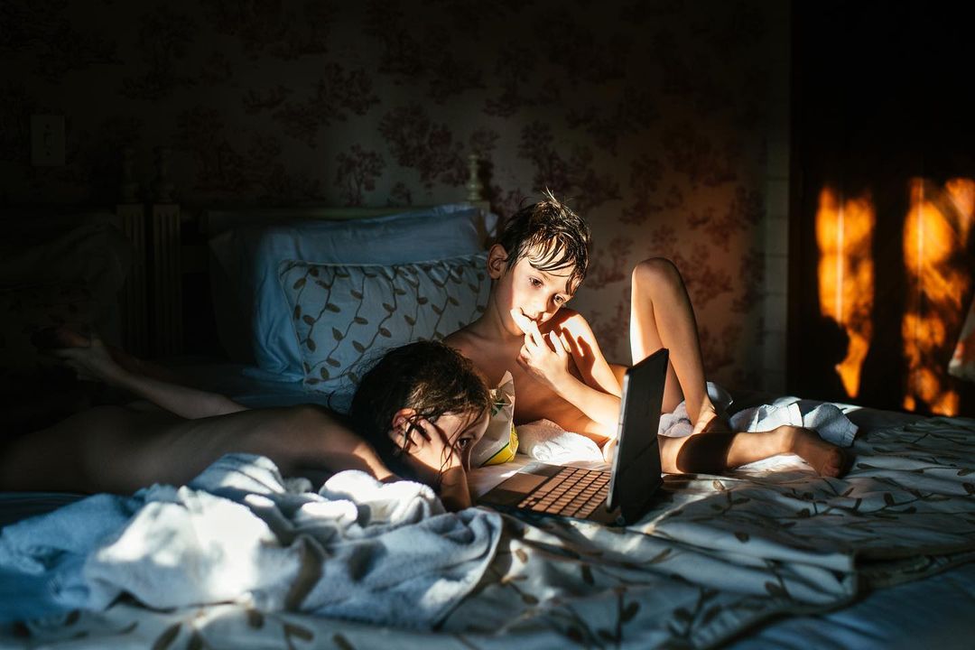 Two children lying on a patterned bed in a dimly lit room while looking at a small illuminated tablet screen.