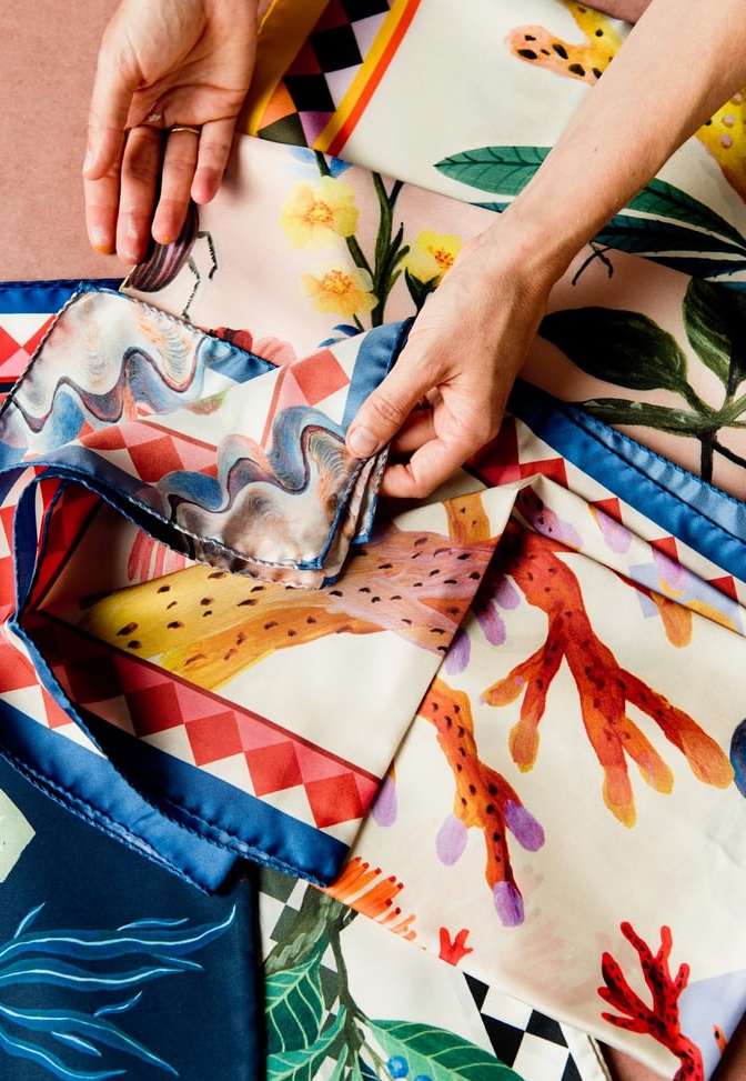 Hands arranging colorful patterned silk scarves with floral and geometric designs on a peach surface, bright lighting, overhead shot.