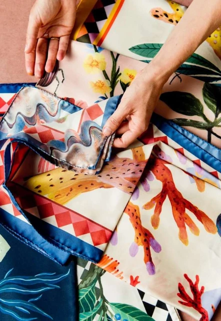 Hands arranging colorful patterned silk scarves with floral and geometric designs on a peach surface, bright lighting, overhead shot.