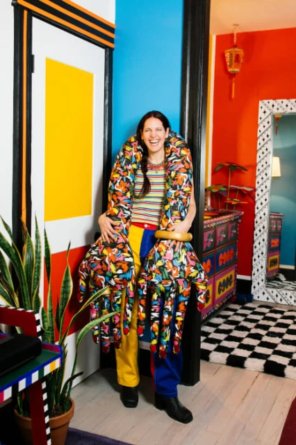 Smiling woman in colourful clothes poses indoors with a jacket made of many hands, next to a colourfully painted door and plant.