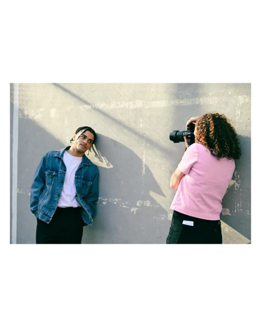 A photographer takes a photo of an adult male model leaning against a weathered concrete wall outdoors in daylight, wearing casual clothing.