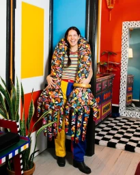 Smiling woman wearing giant patterned sculptural hands and colourful clothing in a brightly painted interior room