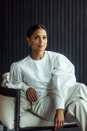 Portrait of a woman wearing a white long-sleeve blouse and light-coloured trousers, seated in an armchair against a dark vertical-slat wall.