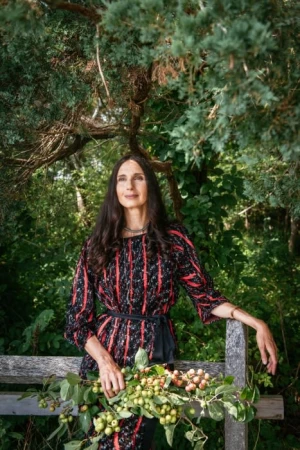 Woman in patterned red and black dress holding branches with green fruit outdoors by a wooden fence