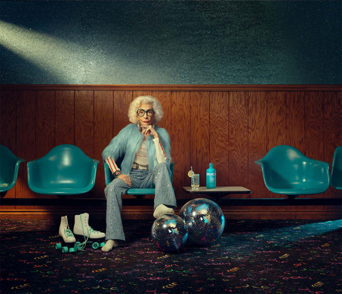Elegant elderly woman with white hair and glasses sitting in a retro teal chair in an old bowling alley, wearing a light blue jacket.