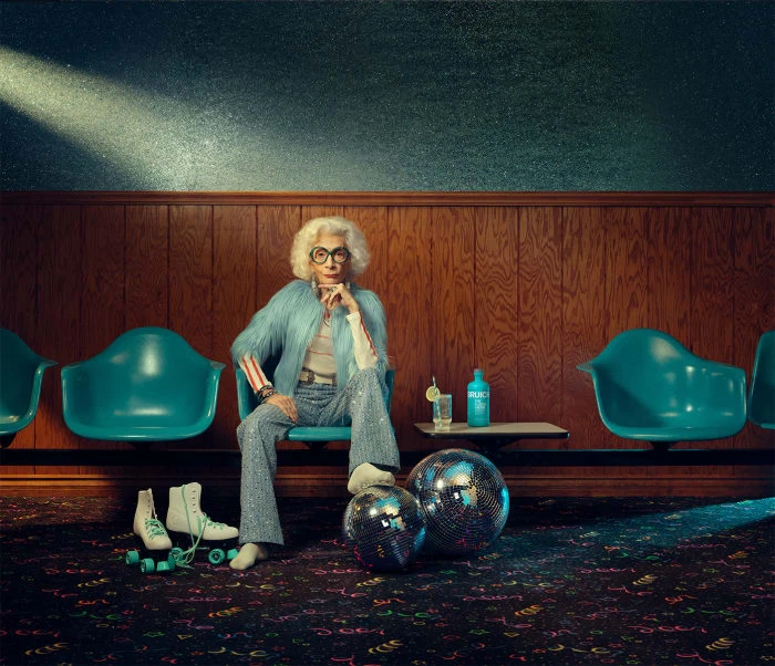 Elegant elderly woman with white hair and glasses sitting in a retro teal chair in an old bowling alley, wearing a light blue jacket.