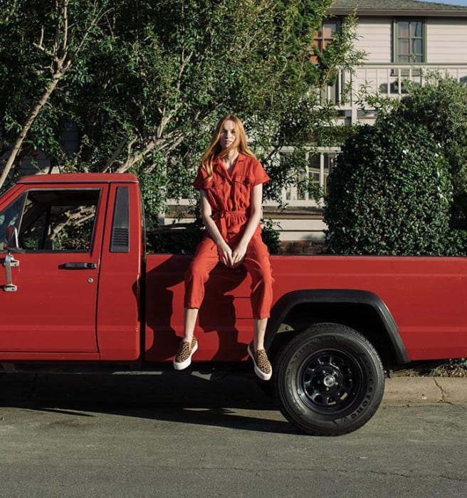 A red-haired woman in a red jumpsuit sits on the back of a red pickup truck on a tree-lined street in front of a house.