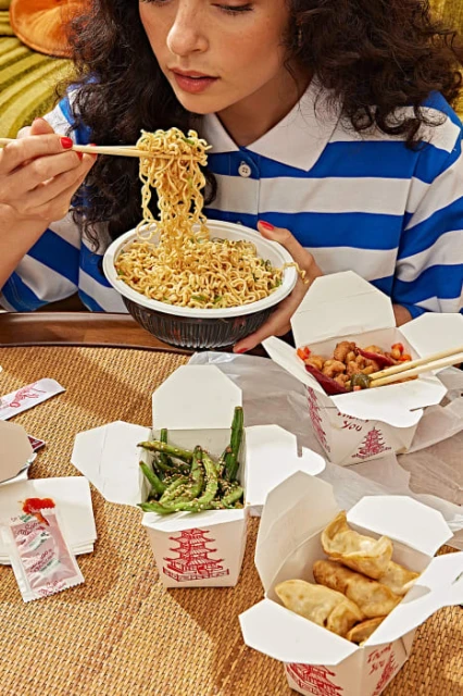 Woman enjoys noodle dish with chopsticks amidst takeout containers on a woven surface, showcasing a casual, food-focused scene.