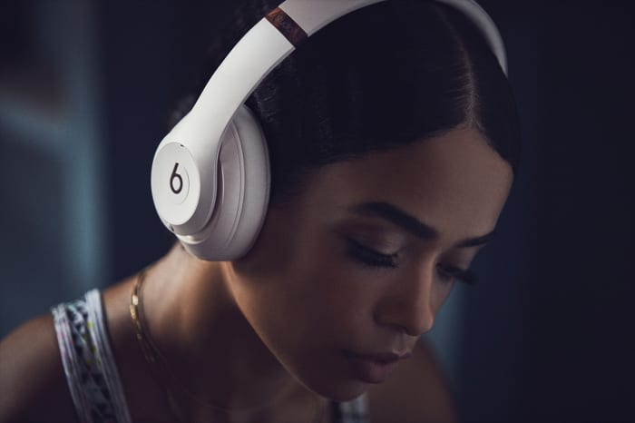 Young woman wearing white Beats headphones with her head down, set against a softly blurred background. Close-up shot, indoor setting.