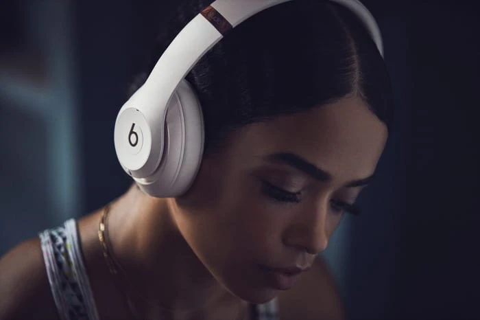 Young woman wearing white Beats headphones with her head down, set against a softly blurred background. Close-up shot, indoor setting.