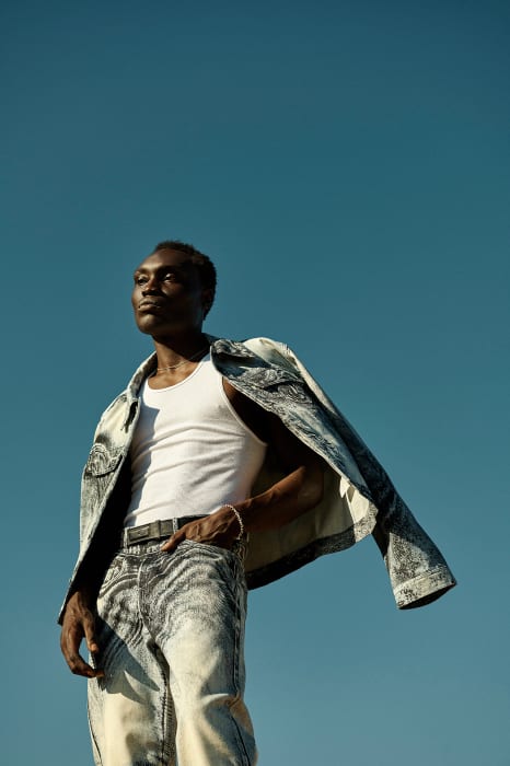 A Black male model poses against a clear blue sky, wearing a white tank top, grey acid wash jeans, and a matching jacket draped over his shoulder.