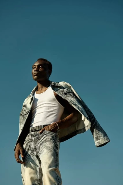 A Black male model poses against a clear blue sky, wearing a white tank top, grey acid wash jeans, and a matching jacket draped over his shoulder.