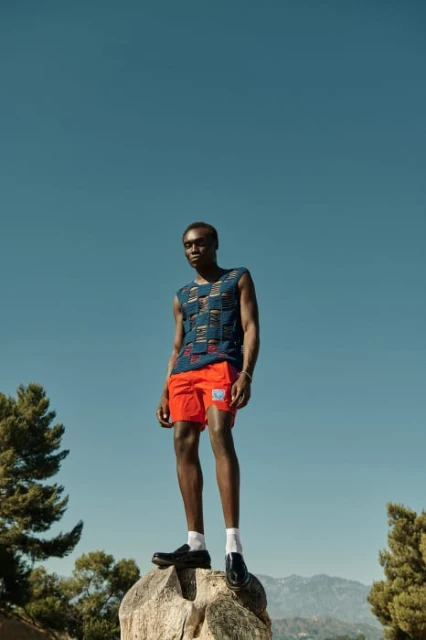 A black male fashion model stands on a rock wearing a patterned tank top and orange shorts in an outdoor daytime fashion editorial.