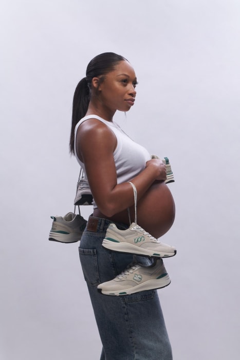 Pregnant woman poses with sneakers hanging around her neck in a side-profile shot against a white backdrop in a studio setting.