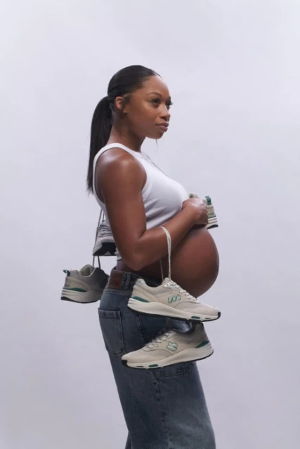 Pregnant woman poses with sneakers hanging around her neck in a side-profile shot against a white backdrop in a studio setting.