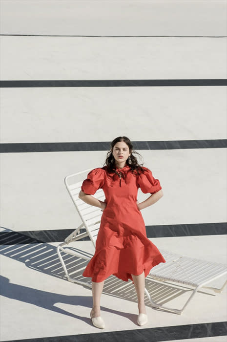 A woman in a red midi dress poses outside next to a white chaise lounge chair and a light colored textured building wall.