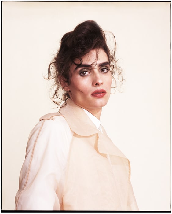 Portrait of an adult woman with curly brown hair wearing a pale, sheer trench coat against a light neutral background in a studio.
