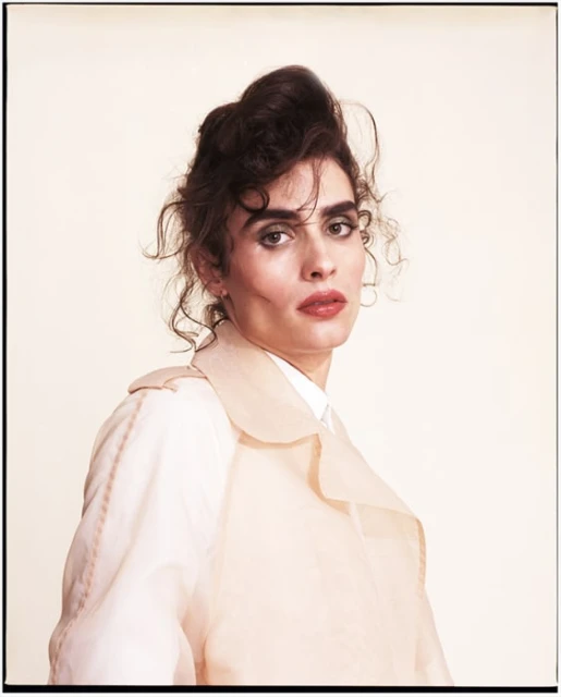 Portrait of an adult woman with curly brown hair wearing a pale, sheer trench coat against a light neutral background in a studio.