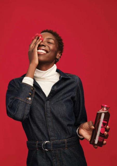 Happy woman in denim jumpsuit holds raspberry drink against a red backdrop in an indoor studio setting with a vibrant and cheerful mood.