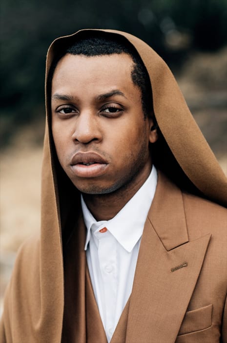 Close-up portrait of a handsome African American male in a caramel-colored suit with a hood, wearing a white shirt, outdoors.
