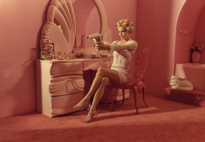 Woman in a pink bathroom sitting in a chair and aiming a hair dryer like a gun. She's wearing a short white dress and clear high heels.