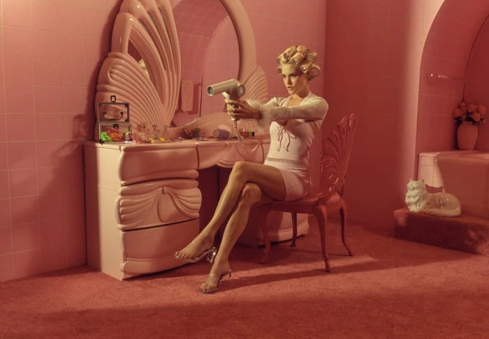 Woman in a pink bathroom sitting in a chair and aiming a hair dryer like a gun. She's wearing a short white dress and clear high heels.