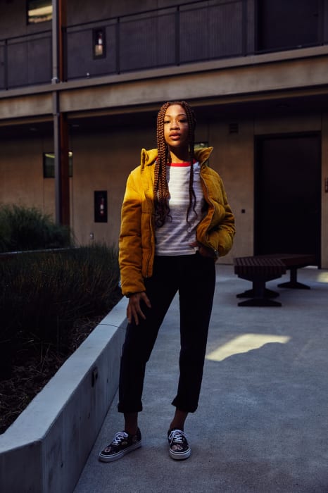 Young woman with braids stands confidently outdoors in a yellow puffer jacket, black pants, and patterned sneakers in front of a building.