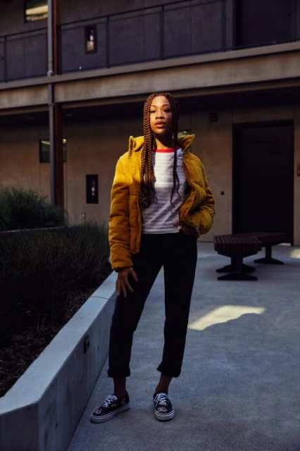 Young woman with braids stands confidently outdoors in a yellow puffer jacket, black pants, and patterned sneakers in front of a building.