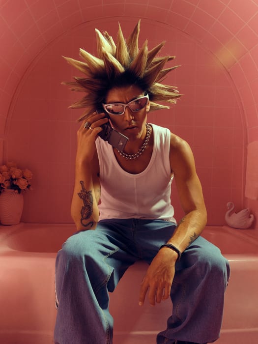 Stylish person with spiky hair sits on a pink bathtub while talking on a cell phone and wearing sunglasses, jewelry, and a tank top.