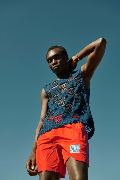 Black man wearing a blue knit tank top and bright orange shorts against a clear blue sky background
