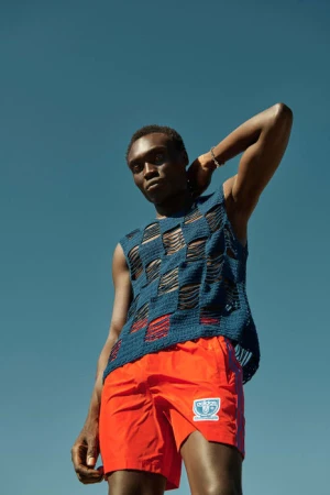 Black man wearing a blue knit tank top and bright orange shorts against a clear blue sky background