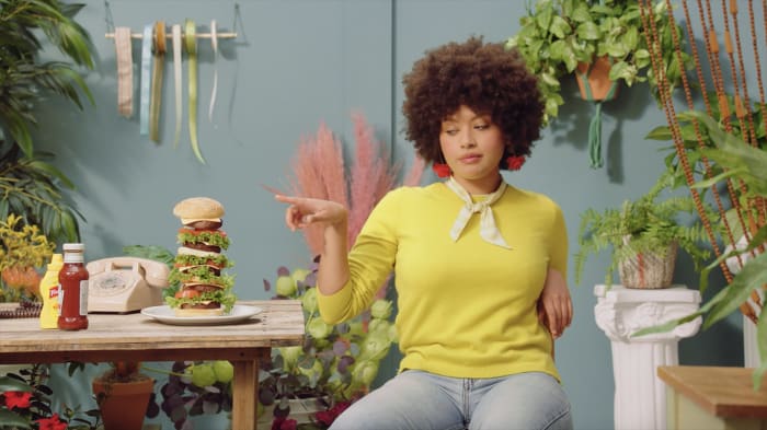 Woman in yellow shirt points to tall burger on table surrounded by indoor plants, vintage phone, condiments and a light blue wall.