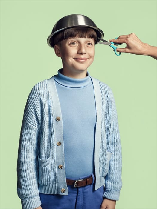 Boy with bowl haircut smiling while someone cuts his hair with scissors against a light green background, creative hair styling.