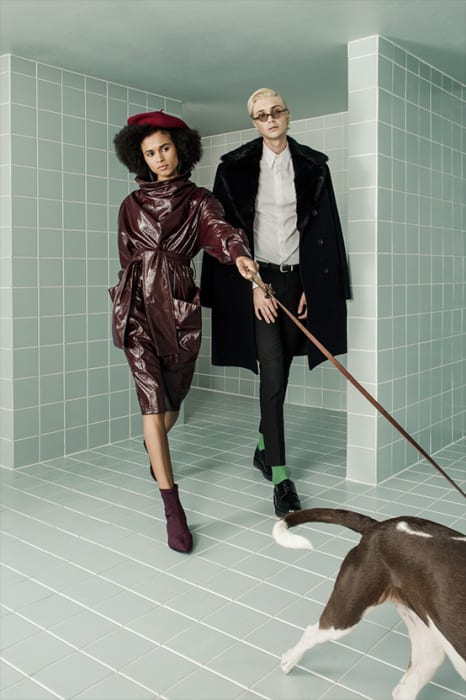 Two fashion models pose indoors in a tiled room with a dog on a leash. One woman wears a burgundy dress, the other a black coat.