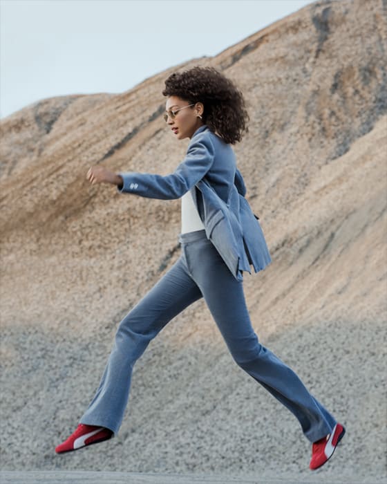 A woman leaps in a blue suit and red sneakers against a rock pile, blending fashion with a natural, rugged backdrop.
