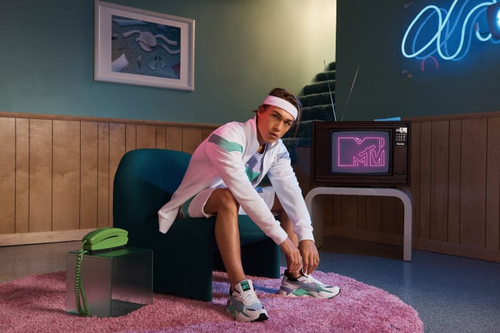 Man sits in teal chair in a retro-styled room, lacing up sneakers, next to a landline phone, with a vintage TV in the background.