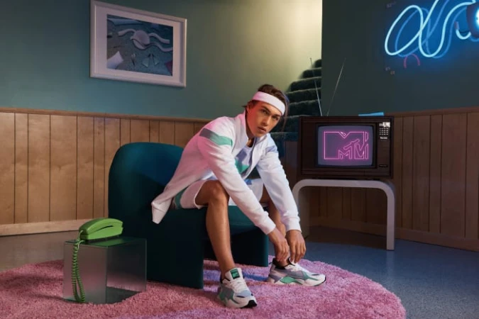 Man sits in teal chair in a retro-styled room, lacing up sneakers, next to a landline phone, with a vintage TV in the background.