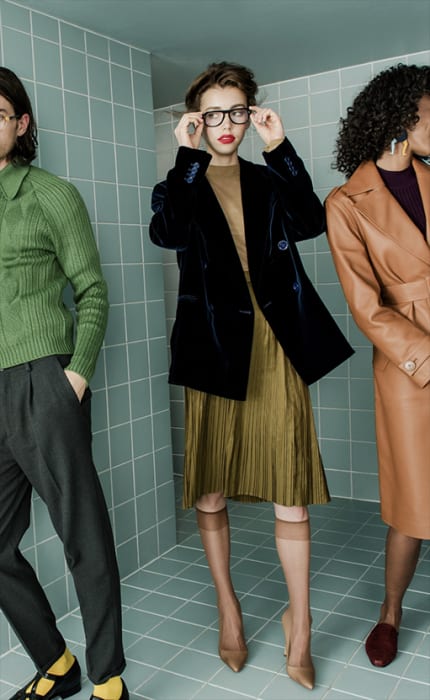 Three fashion models pose indoors against a blue-tiled wall, showcasing a green knit top, a blue velvet blazer, and a tan trench coat.